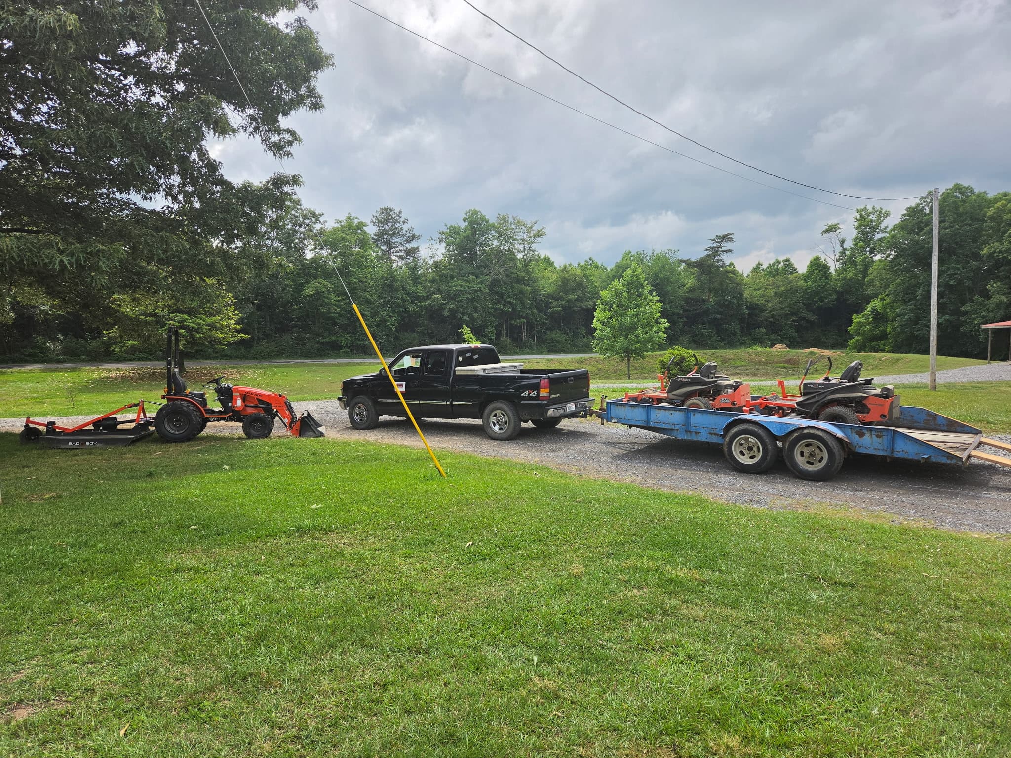 Big Boys Landscaping rig — compact tractor with rotary cutter, pickup, and trailer loaded with zero-turn mowers